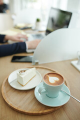 Cup of cappuccino and silice of cake on cafe table of businesswoman working on laptop