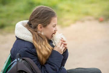 A teenage schoolgirl girl is sitting on a bench with a backpack in a medical mask and eating shawarma