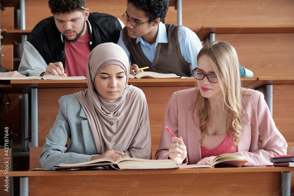 Poster two intercultural female students of university sitting by desk in lecture hall and looking through 
