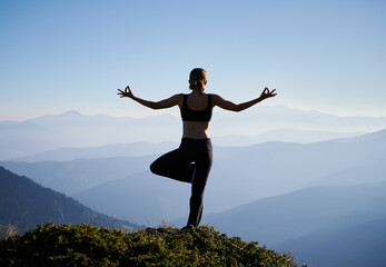 Silhouette of woman practicing yoga on background of evening mountains. Meditating female is balancing on one leg after sunset. Concept of yoga.