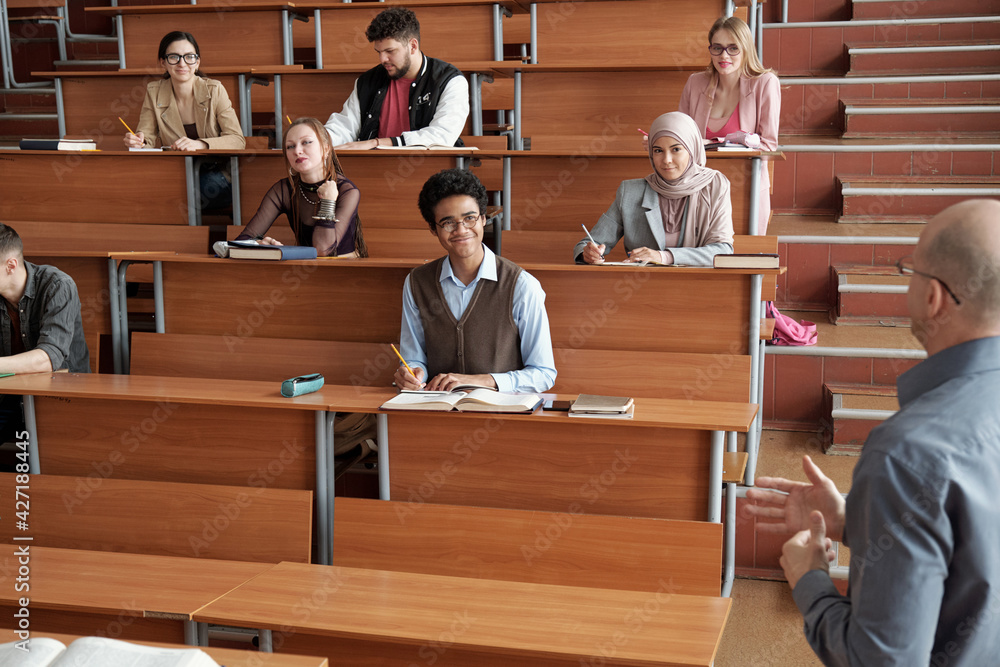 Poster happy intercultural university students sitting by long wooden desks in lecture hall, making notes a
