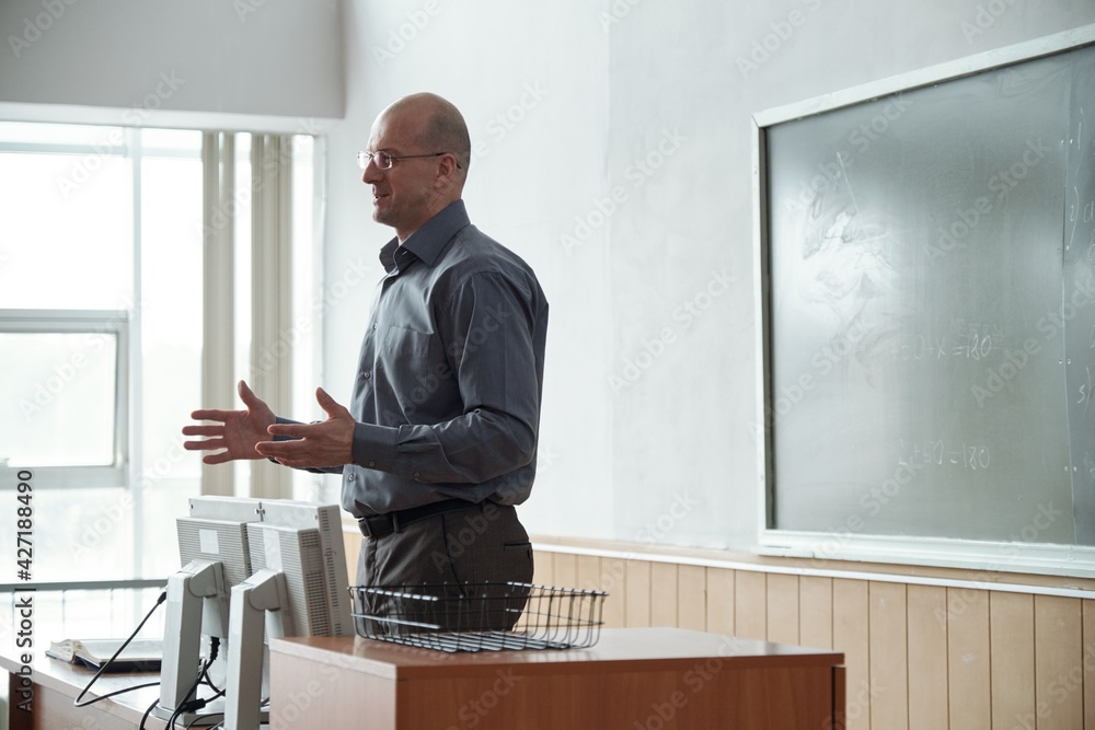 Poster confident bald mature professor in smart casualwear standing by desk in front of audience and explai