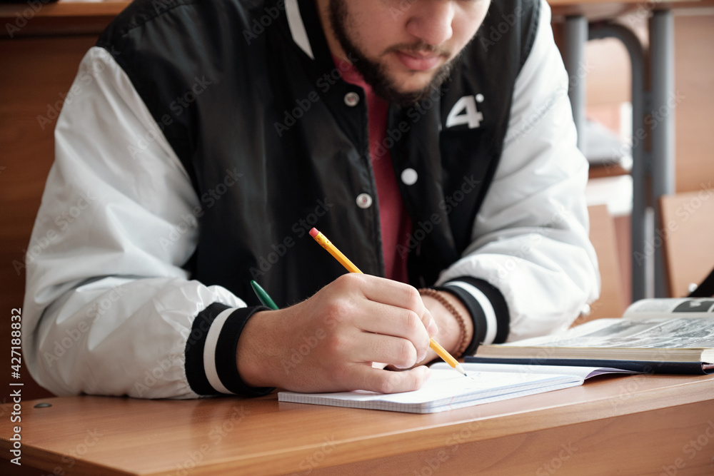 Poster Hands of bearded guy in casualwear bending over desk and making notes or drawing sketch in copybook during lecture in auditorium of university