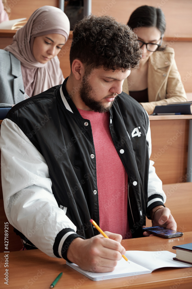 Poster Serious guy in casualwear sitting by desk and making notes in copybook while scrolling in smartphone during individual work at lesson