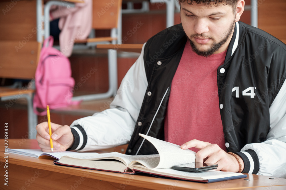 Poster young male student cheating while sitting by desk, making notes in copybook and looking at prompt on