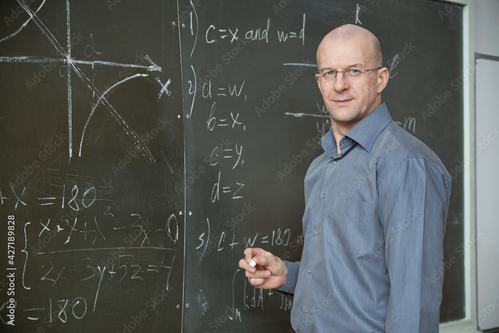 Poster mature bald professor in smart casualwear holding piece of chalk while standing by blackboard in cla