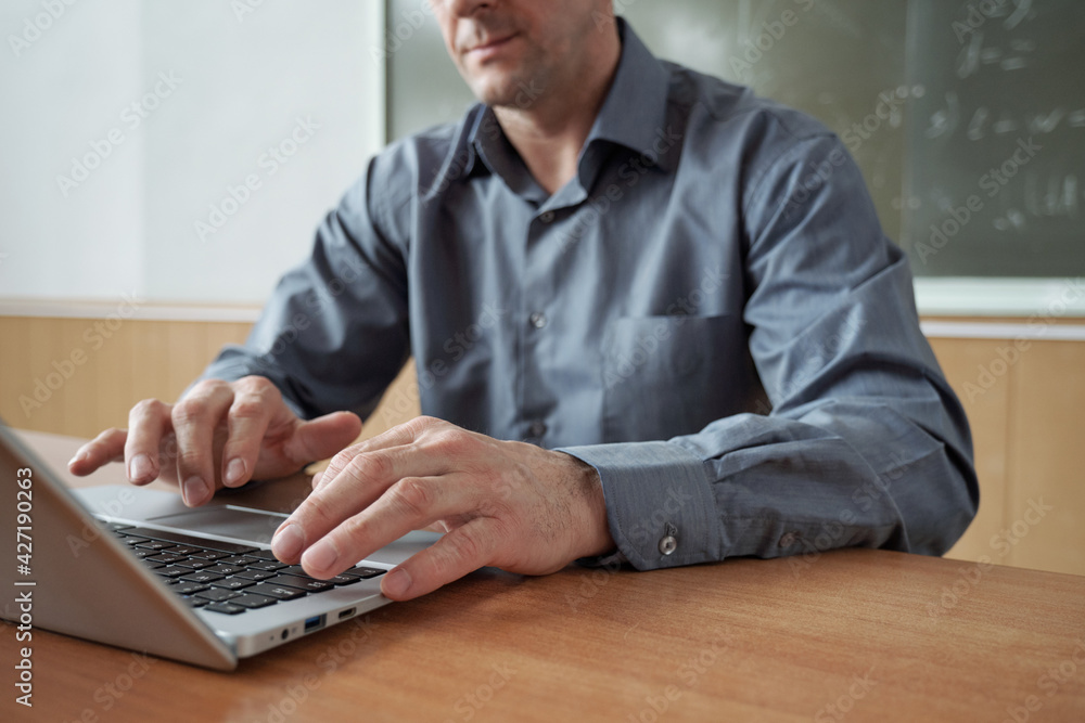 Poster hands of mature man in grey shirt touching buttons of laptop keypad while sitting by desk against bl