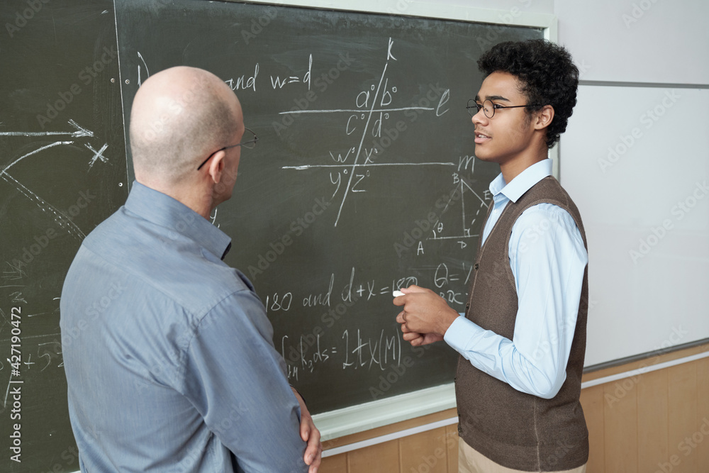 Poster mature bald professor and young mixed-race student standing by blackboard and discussing solution of