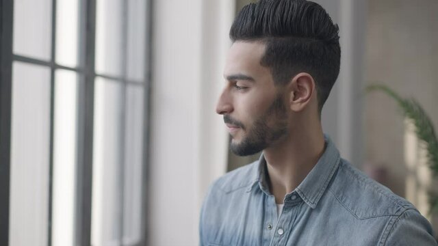 Portrait of handsome smiling young Middle Eastern man looking at camera and looking out the window. Positive confident millennial indoors at home in the morning