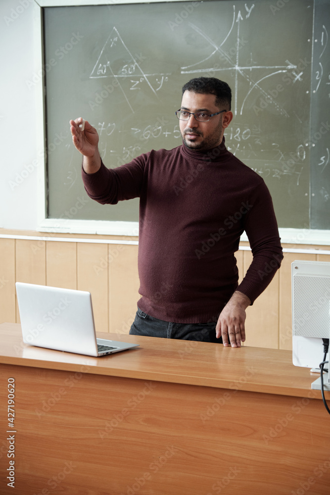 Poster mixed-race male in casualwear holding piece of chalk while standing between desk and blackboard and 