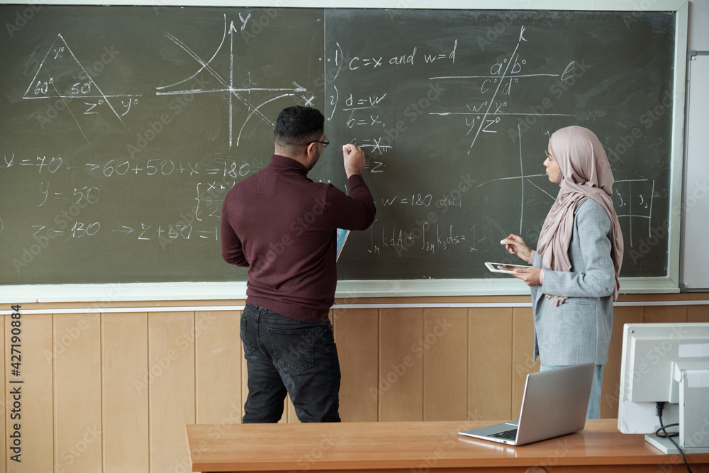 Poster male professor and young female student in hijab standing by blackboard and solving equation at less