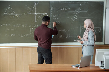 Male professor and young female student in hijab standing by blackboard and solving equation at...
