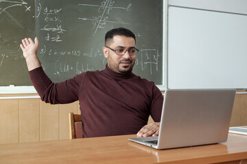 Mixed-race male teacher in casualwear pointing at solution of equation on blackboard while sitting...