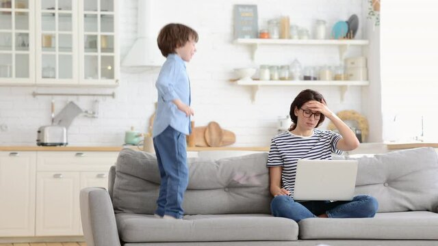 Irritated mother sitting on couch working on laptop at home, distracted from noise and touching her temple, hyperactive little kid jumping on sofa and drawing attention. Tired mom can't concentrate.