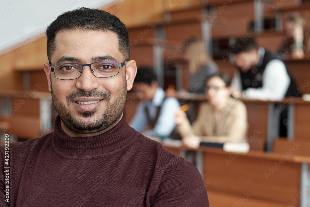 Poster smiling male teacher in eyeglasses and maroon pullover standing in front of camera against group of 