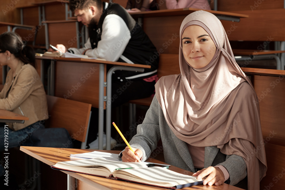 Poster young smiling female student in hijab sitting by desk at lesson in lecture hall and looking you whil