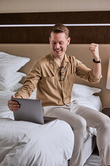 Positive businessman sitting on bed using laptop,joyful and raising fist, happy and satisfied with result