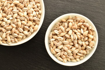 Organic uncooked pearl barley in a white dish on a slate board, close-up, top view.
