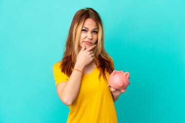 Teenager blonde girl holding a piggybank over isolated blue background thinking