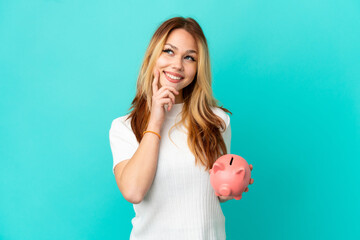 Teenager blonde girl holding a piggybank over isolated blue background thinking an idea while looking up