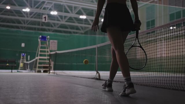 confident sporty woman is walking over tennis court, holding tennis racquet and throwing ball, view on slender trained legs