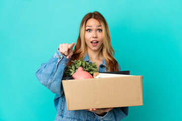 Teenager blonde girl making a move while picking up a box full of things surprised and pointing front