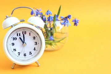 White alarm clock on the background of a bouquet in a glass vase, yellow background, a place for text-the concept of the arrival of spring days