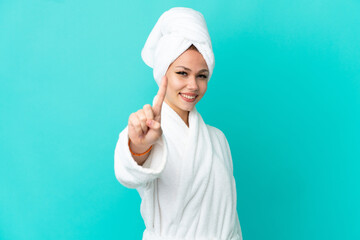 Teenager blonde girl in a bathrobe over isolated blue background showing and lifting a finger