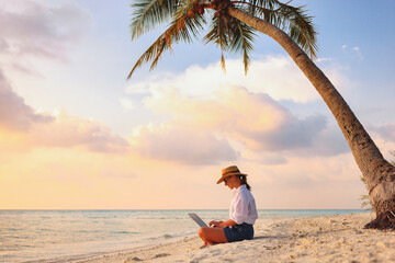 Young female freelancer wearing straw hat working on laptop while sitting on tropical beach at...
