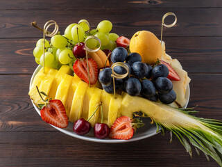 Party plate with fresh fruit and berries. Pineapple, blue and green grapes, strawberries, cherries, melons and apricots. On a wooden background. Fruit for the festive table. Copy space. Rustic style.