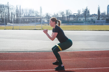 Fototapeta premium Fitness and sport concept, outdoor workout. Young sportive woman work out with the help of fitness rubber bands on the red track.
