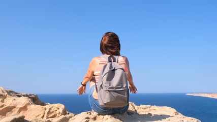 young happy woman with a backpack sits on a rock with her hands raised and looks at the valley below. The concept of freedom and ease.