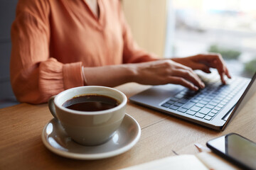 Close up shot of unrecognizable young businesswoman using laptop while enjoying work at cafe table with cup of coffee, copy space