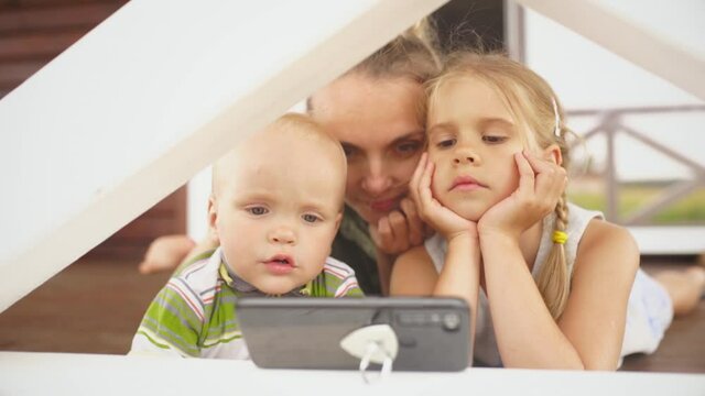 Two little children and their smiling blond mom looking at photos in smartphone 