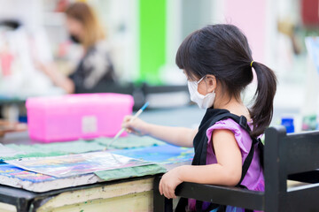 Selective focus. Rear back  girl wear face mask is in class. Student is studying art. Child paints the water with paintbrush in his right hand. Kid make crafts on canvas. Baby wearing a black apron.