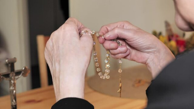 Christian old woman prays with rosary. Dressed in black. In her hands she holds the rosary beads and the cross. In the background, there is a cross on the table. Shot by the shoulder.