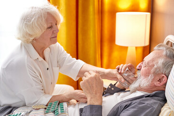 aged female concerned with health condition of husband, checking fever temperature lying on bed