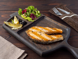 Grilled salmon steak on a wooden board. Serve salmon steak in the restaurant with lemon, olives and green salad.  Juicy fried delicious fish. Space for text. Rustic style. Copy space.