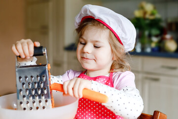 Cute little preschool girl grate carrots for baking carrot cake or cookies. Happy toddler child in...