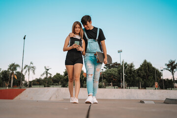 Fototapeta premium Young couple walking, carrying a skateboard and looking at their mobile phone in the park.