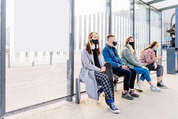 Group of young male and females people im protective masks sitting in line at bus stop. Keeping safe social distance. Hipsterh man and women outdoor waiting for transport.