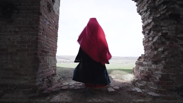 Woman in red dress and headscarf stands in an abandoned building near the doorway. Wind blows and the fabric flies in an abandoned church  