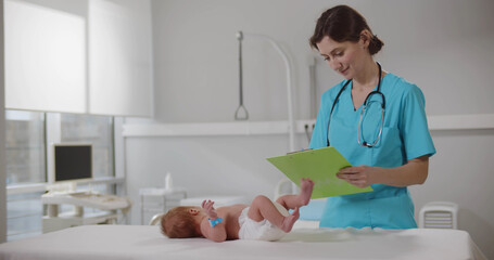 Young nurse writing down examination data of cute newborn baby lying on table