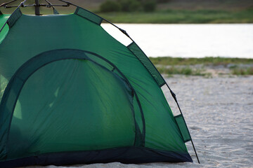 Green tent on sandy beach against river background. Camping, trekking, hiking