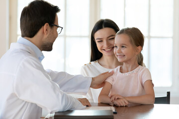 Caring male doctor in white medical uniform consult happy little girl patient visiting hospital with mom. Man pediatrician or GP comfort smiling small kid child at consultation in clinic with mother.