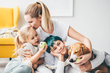 Young mother with pretty little daughter eating apple and teenage son have fun with beagle in light...