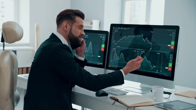 Confident young man in shirt and tie analyzing data on the stock market and talking on the phone while working in the office