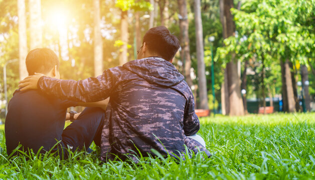 two asian man friends sitting on green grass in the park, encouraging, comforting his friend and looking in the same direction