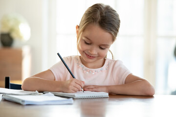 Cute smart little 7s girl child sit at table at home write in exercise book prepare homework alone. Happy small kid do task assignment, handwrite in notebook, study for school. Education concept.