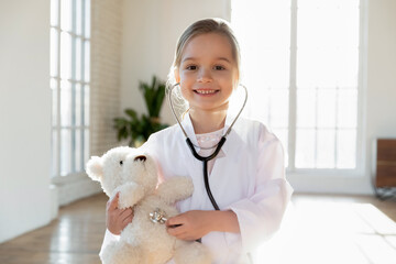 Portrait of smiling little Caucasian girl child in white medical uniform cure stuffed teddy bear play hospital. Happy small kid act as doctor examine toy in vet clinic. Career, healthcare concept. - obrazy, fototapety, plakaty